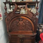 Highly ornate dark-wood secretary cabinet featuring carved winged figures, a central cherub panel, decorative hardware, drawers, and lower storage.