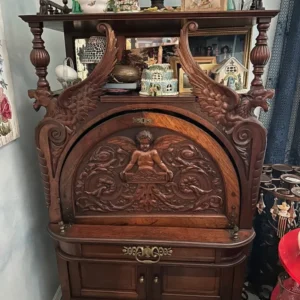 Highly ornate dark-wood secretary cabinet featuring carved winged figures, a central cherub panel, decorative hardware, drawers, and lower storage.
