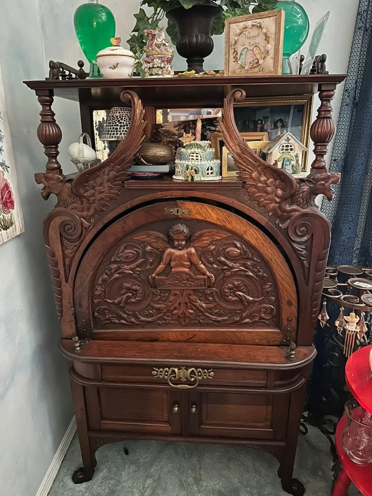 Highly ornate dark-wood secretary cabinet featuring carved winged figures, a central cherub panel, decorative hardware, drawers, and lower storage.
