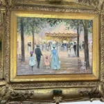 Colorful framed street scene depicting finely dressed figures near an outdoor pavilion or café in an elaborate gilt frame.