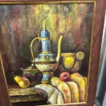 Framed still life featuring a decorative wine vessel, goblets, fruit, and bread against a dark background.